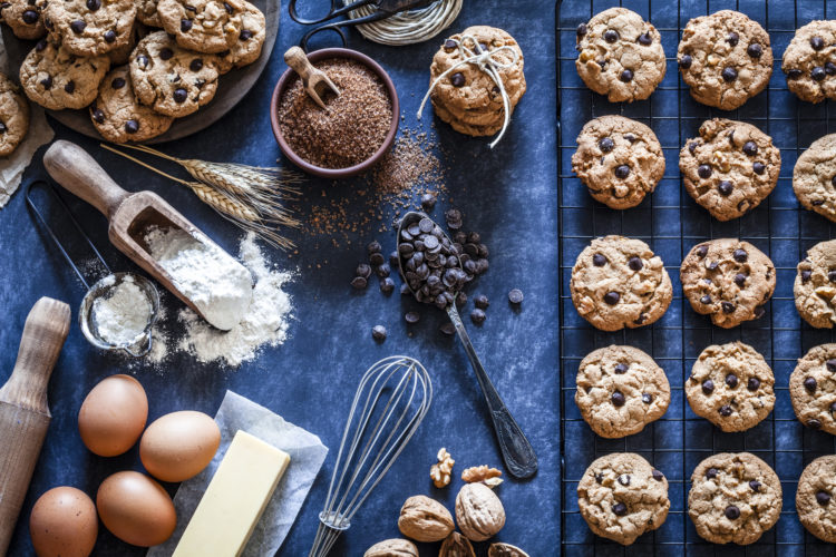 Top view of a dark kitchen table filled with chocolate chips cookies and ingredients for preparing and baking cookies. Some baked cookies are on a cooling rack and on a wooden plate. Ingredients includes are flour, eggs, chocolate chips, brown sugar and butter. Some kitchen utensils complete the composition.